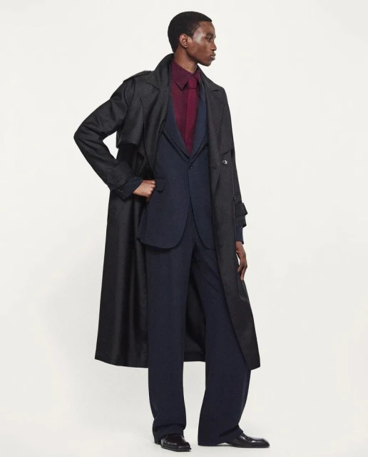 An adult Black male poses in a full-length shot, wearing a dark overcoat, navy suit, and burgundy shirt, set against a neutral backdrop.