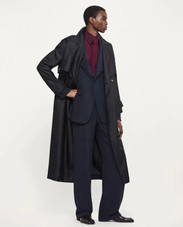 An adult Black male poses in a full-length shot, wearing a dark overcoat, navy suit, and burgundy shirt, set against a neutral backdrop.
