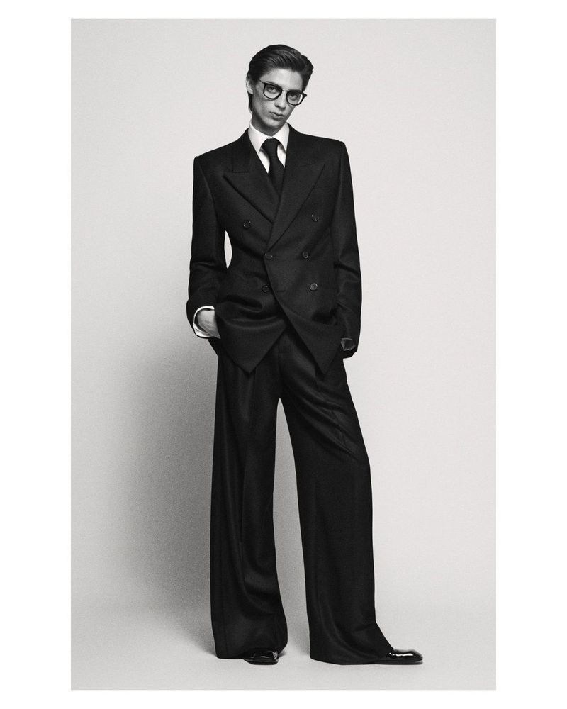 Adult male model in dark double-breasted suit, glasses, and wide-leg pants poses in a studio with hands in pockets. Black and white fashion shot.