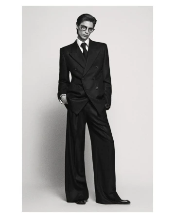 Adult male model in dark double-breasted suit, glasses, and wide-leg pants poses in a studio with hands in pockets. Black and white fashion shot.