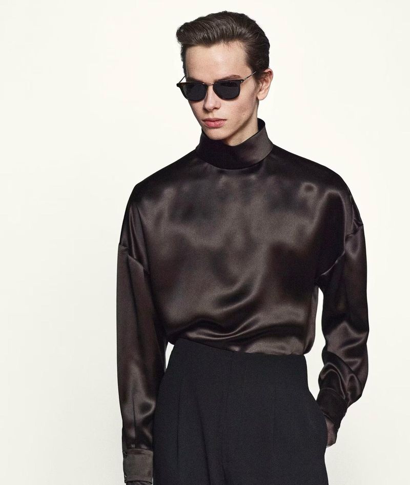 Male model wearing a brown satin long-sleeve blouse and black pants against a white background in a fashion studio shot.