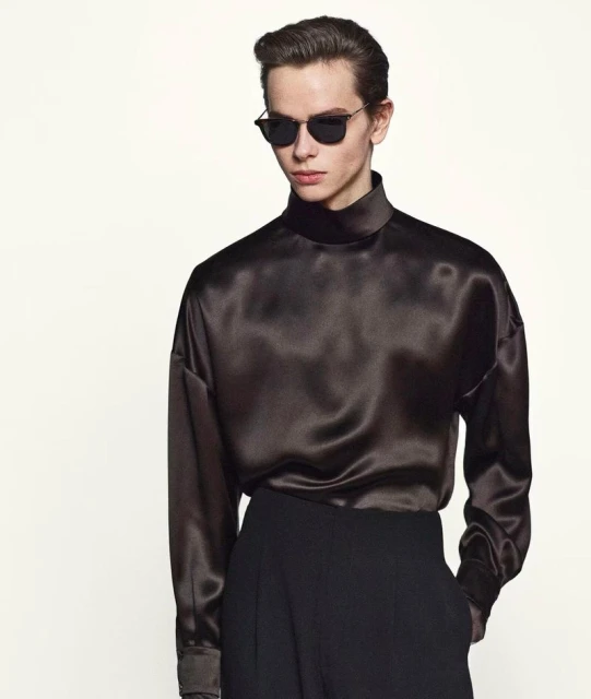 Male model wearing a brown satin long-sleeve blouse and black pants against a white background in a fashion studio shot.