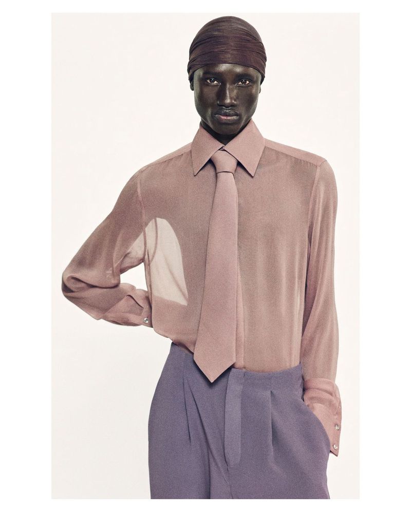 A dark-skinned adult man in a semi-sheer mauve button-down shirt, mauve tie, and lilac-purple dress pants stands in a light studio.