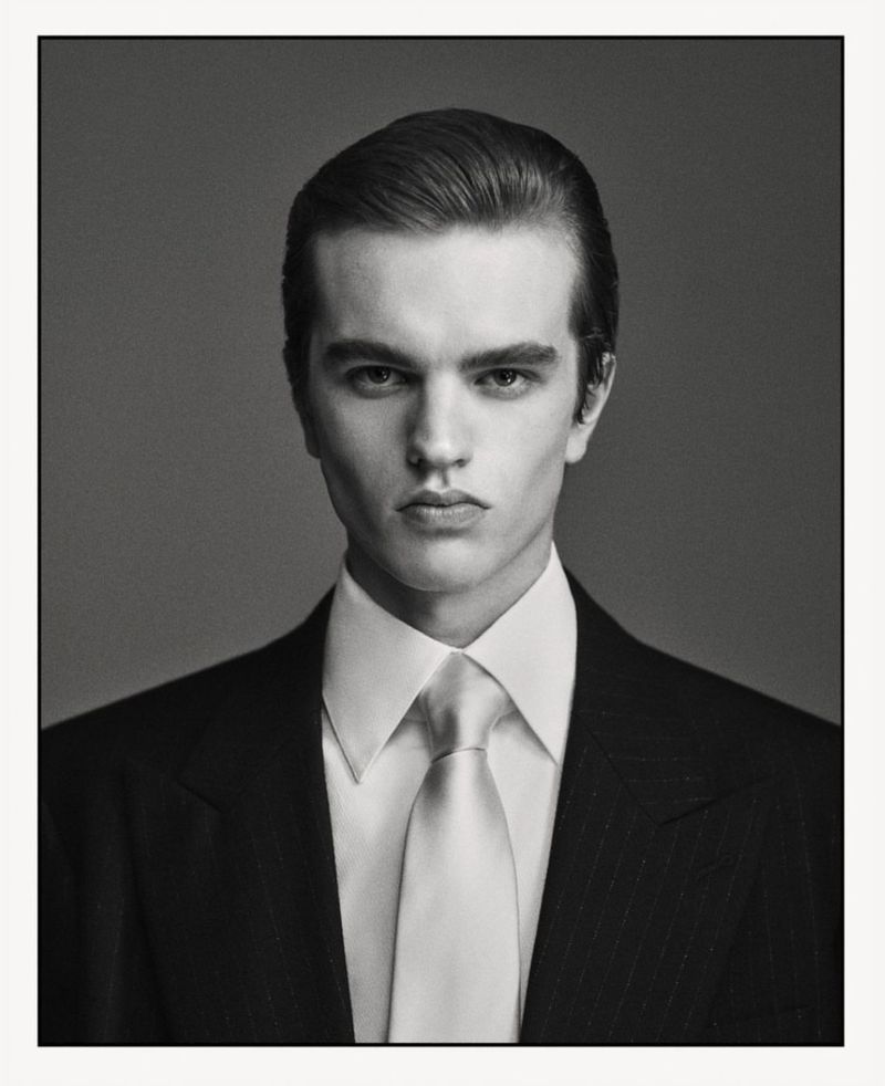 Headshot of a young adult man wearing a suit and tie. He is looking directly at the camera in a studio setting with a grey background.