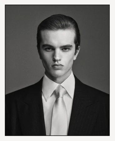 Headshot of a young adult man wearing a suit and tie. He is looking directly at the camera in a studio setting with a grey background.