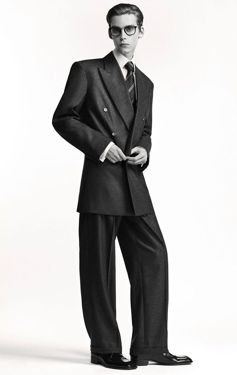 Male model wearing black eyeglasses and a charcoal double-breasted suit against a pure white studio backdrop.