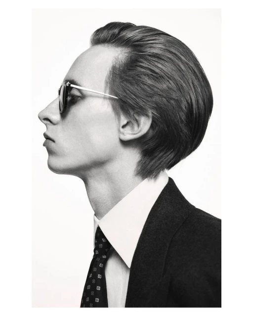 Headshot of a fair-skinned adult male in a dark suit, white collared shirt, and patterned tie, wearing sunglasses against a bright background.