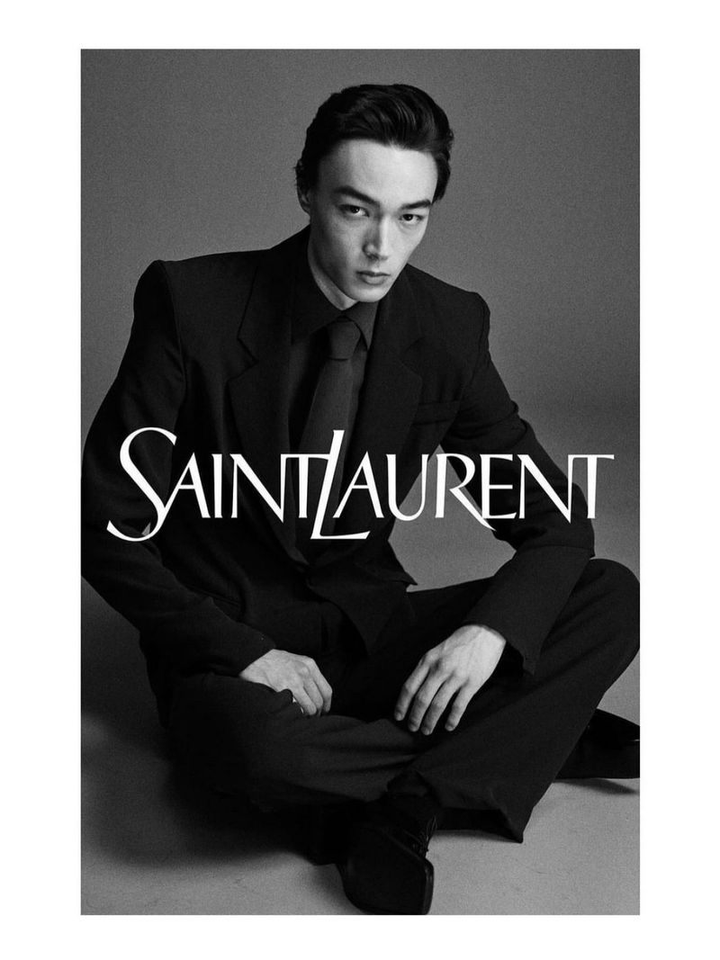 Black-and-white portrait of a young man in a dark suit and tie, seated against a neutral backdrop with 'SAINT LAURENT' logo.