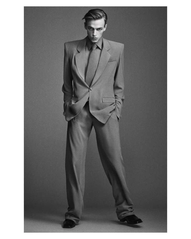 Monochrome portrait of adult man in full suit with necktie standing in front of a gray backdrop in a full length view indoors.