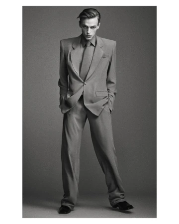 Model wearing an oversized grey tailored suit with wide padded shoulders and matching tie against a seamless grey backdrop.