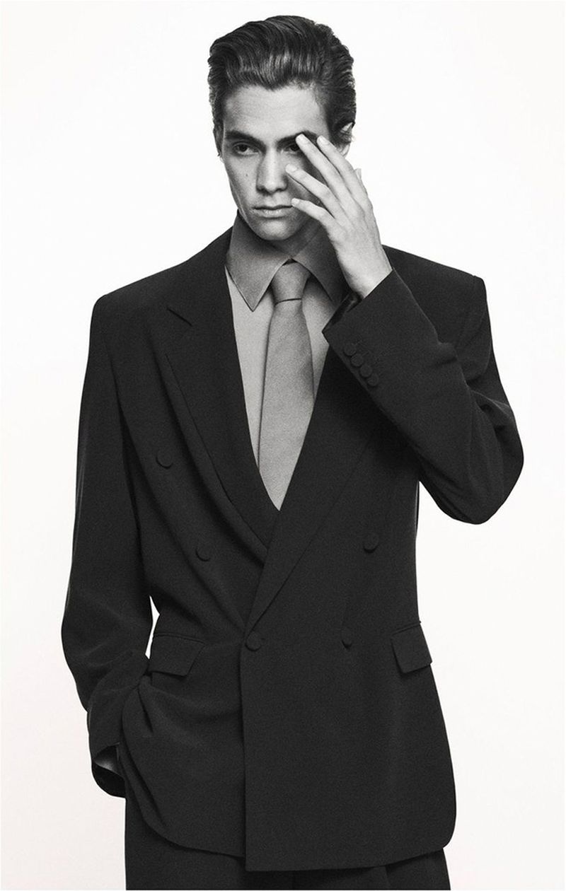 Monochrome portrait of a man in a suit with his hand near his face, posing in a photo studio against a white backdrop.