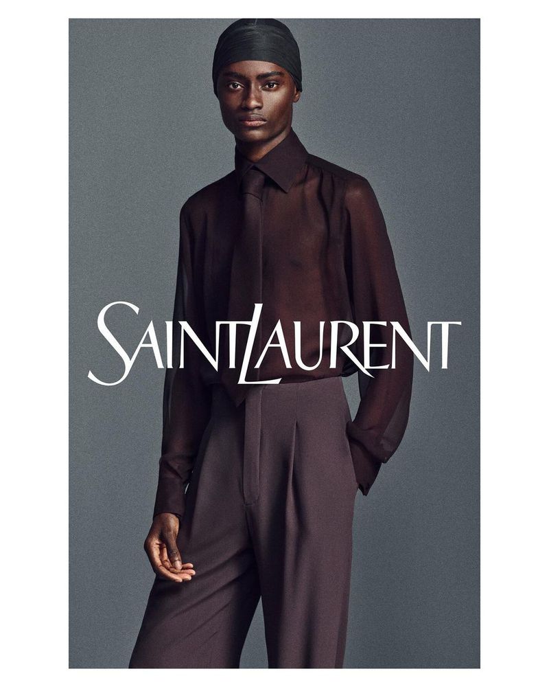 Dark-skinned adult fashion model poses in a sheer dark top and high-waisted trousers for a studio fashion shot, promoting Saint Laurent.