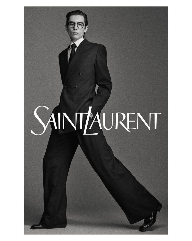 Black and white fashion portrait of a slender male model in a dark suit, dress shirt, and tie, posing in front of the Saint Laurent logo.