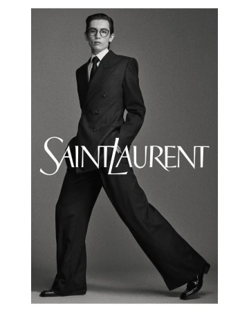 Black and white fashion portrait of a slender male model in a dark suit, dress shirt, and tie, posing in front of the Saint Laurent logo.