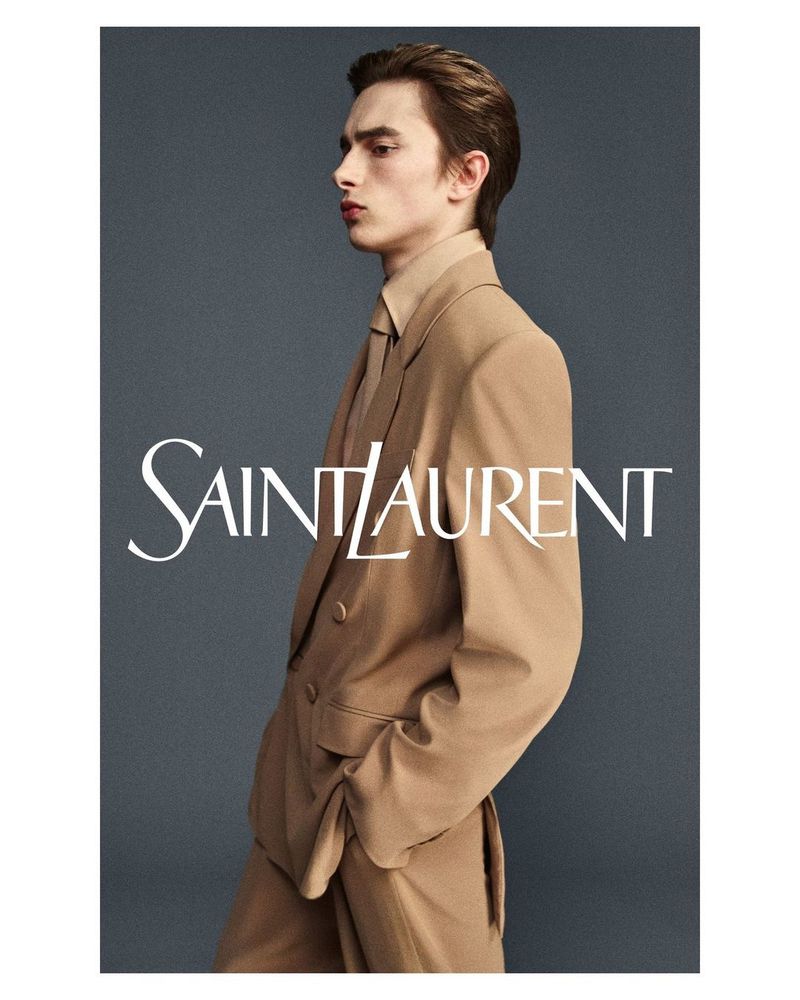 Fashion shot of a young adult male in a tan suit and tie against a grey backdrop with the Saint Laurent logo in white.