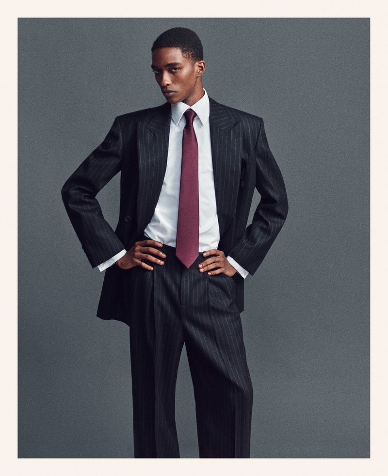 Man poses in pinstripe suit with white shirt and maroon tie in front of a plain grey background. Fashion editorial style.