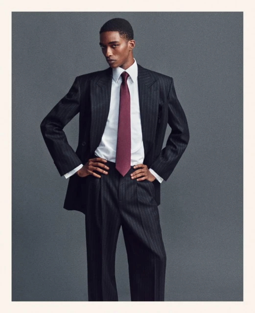 Man poses in pinstripe suit with white shirt and maroon tie in front of a plain grey background. Fashion editorial style.