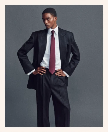 Man poses in pinstripe suit with white shirt and maroon tie in front of a plain grey background. Fashion editorial style.