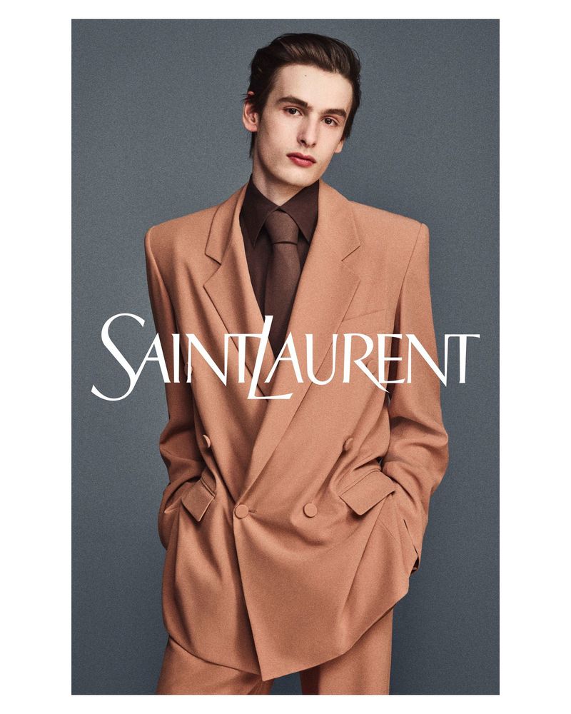 A male model posing in a camel double-breasted suit and brown shirt against a grey seamless background for Saint Laurent.