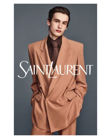 A male model posing in a camel double-breasted suit and brown shirt against a grey seamless background for Saint Laurent.