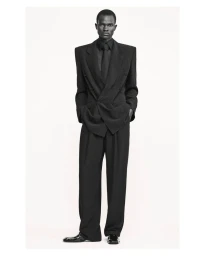 Male model standing in an oversized black double-breasted suit and tie against a plain white studio backdrop.
