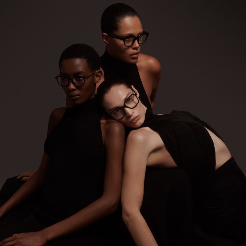 Three female models in black dresses pose in a studio, wearing square glasses. Fashion shot with soft lighting.