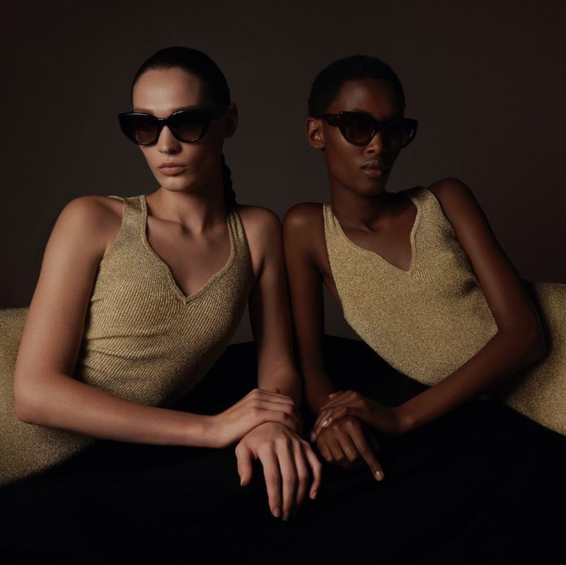 Two women pose indoors wearing sunglasses and gold tops against a dark background, showcasing fashion and style.