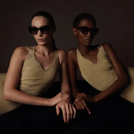 Two women pose indoors wearing sunglasses and gold tops against a dark background, showcasing fashion and style.