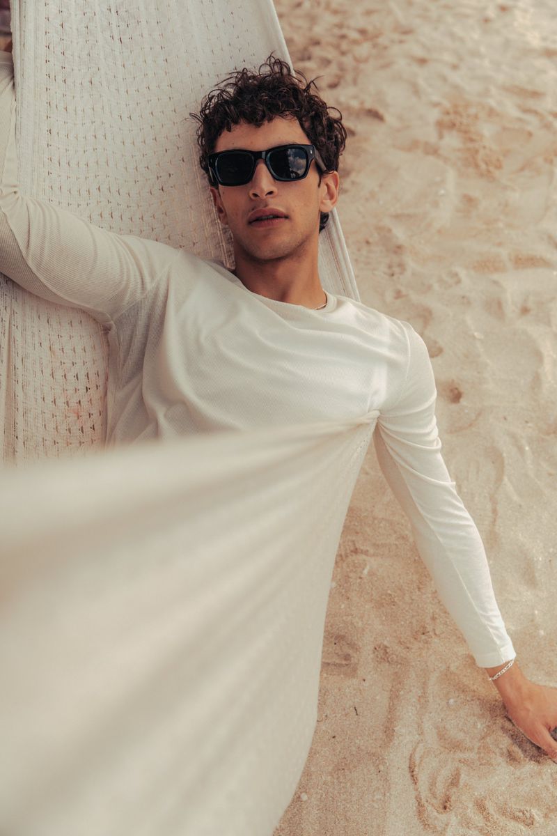 Adult male model in white long-sleeve shirt and black sunglasses reclining in a hammock on a sandy beach with soft lighting.