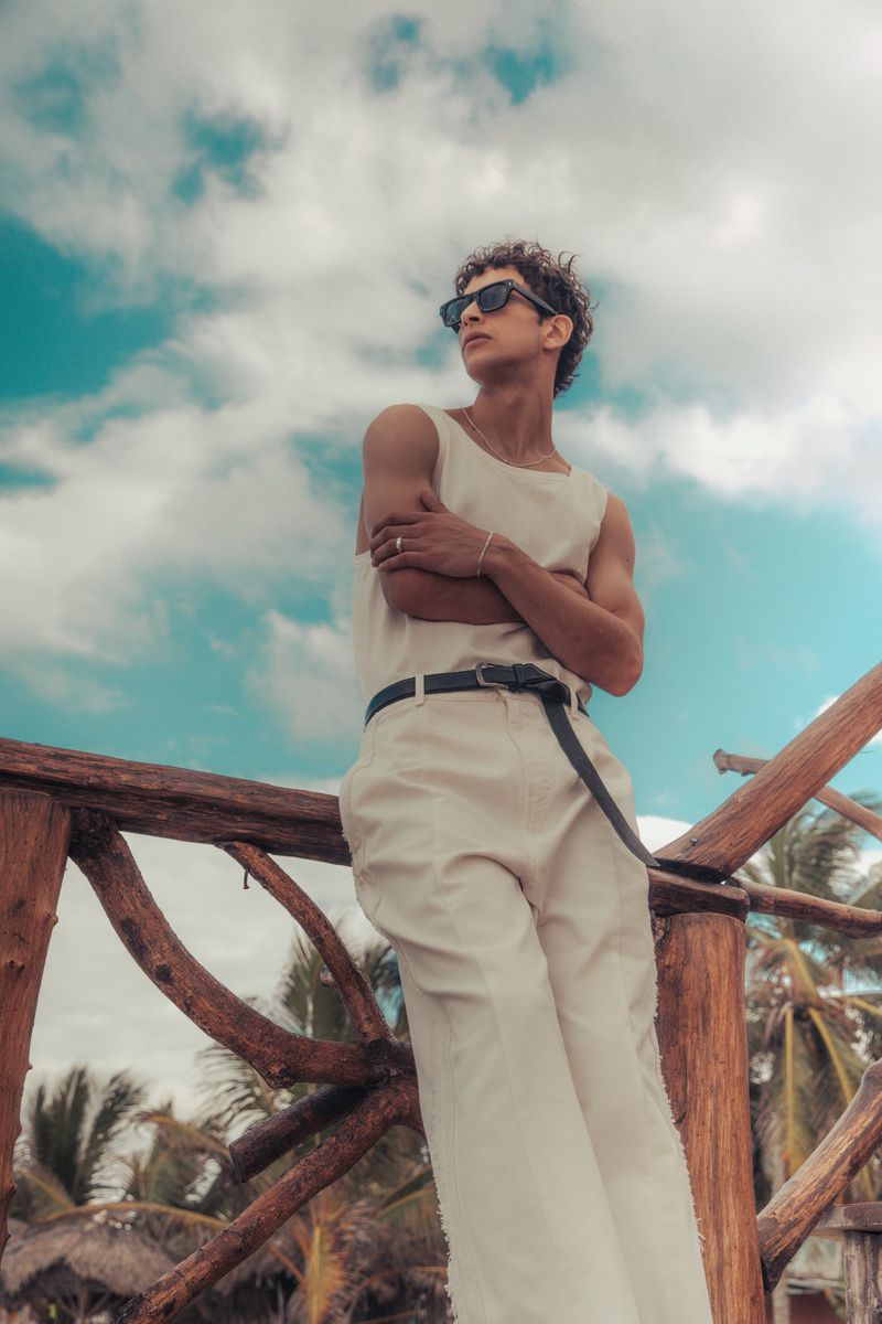 Portrait of adult male in white sleeveless shirt and trousers leaning against a wooden railing with trees and a cloudy sky in the background.