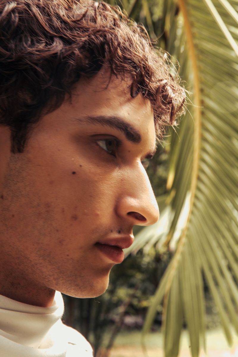 Close-up profile portrait of a young adult male with curly hair near a palm leaf, creating a natural, outdoor aesthetic.