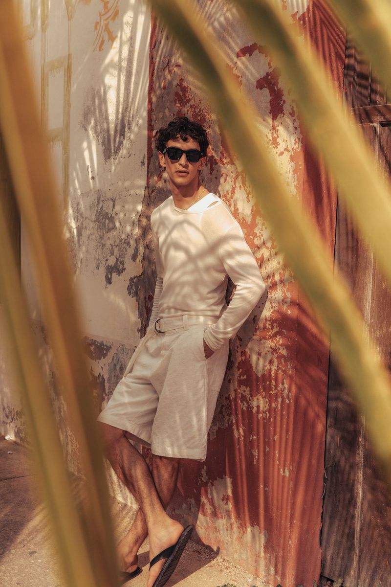 A young man wearing sunglasses, a white long-sleeve shirt, and white shorts poses against a weathered wall with palm fronds in the foreground.