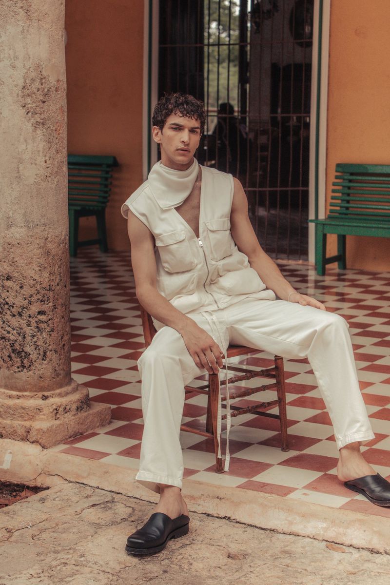 A slender man sits on a wooden chair wearing an off-white zip up vest, a matching top and pants, and black leather shoes.