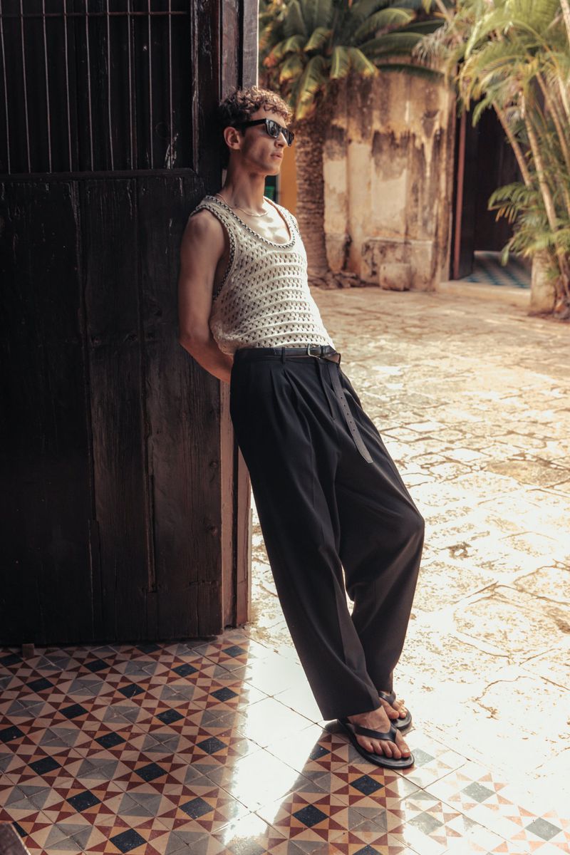 A man in sunglasses and a crochet tank top leans against a weathered door in an outdoor courtyard with patterned tiles and greenery.