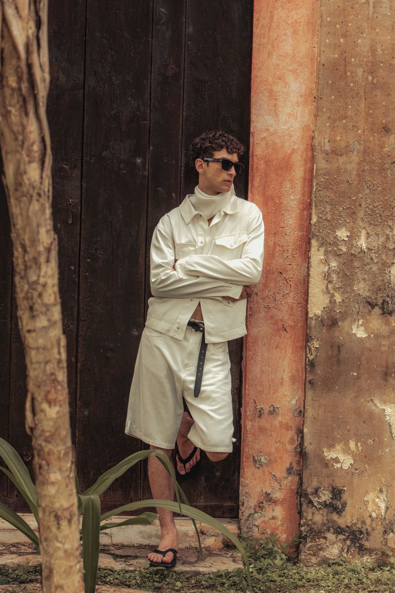 An adult male in a white outfit poses against a weathered wall next to a dark door outdoors, wearing sunglasses and sandals.