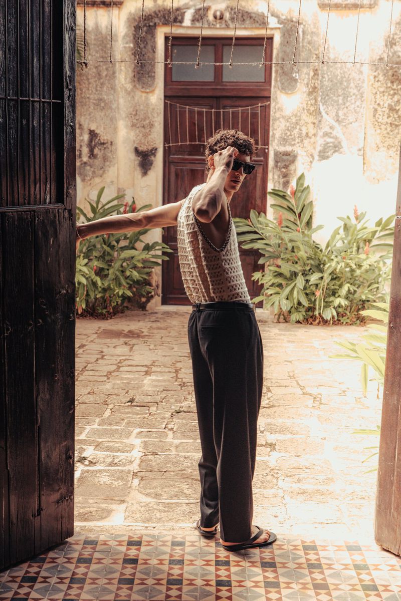 A slender man wearing a crocheted tank top and sunglasses stands in an open doorway in a vintage outdoor patio with tropical plants.