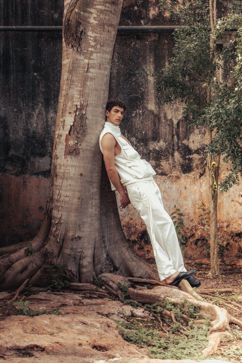 Male fashion model poses in sleeveless top and trousers leaning against large tree, outdoors with textured wall and foliage backdrop.