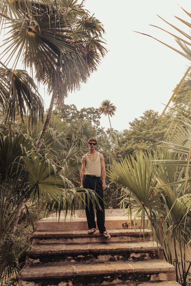 A full shot of a young adult man standing on stone steps, surrounded by tropical foliage in a sunny outdoor environment.