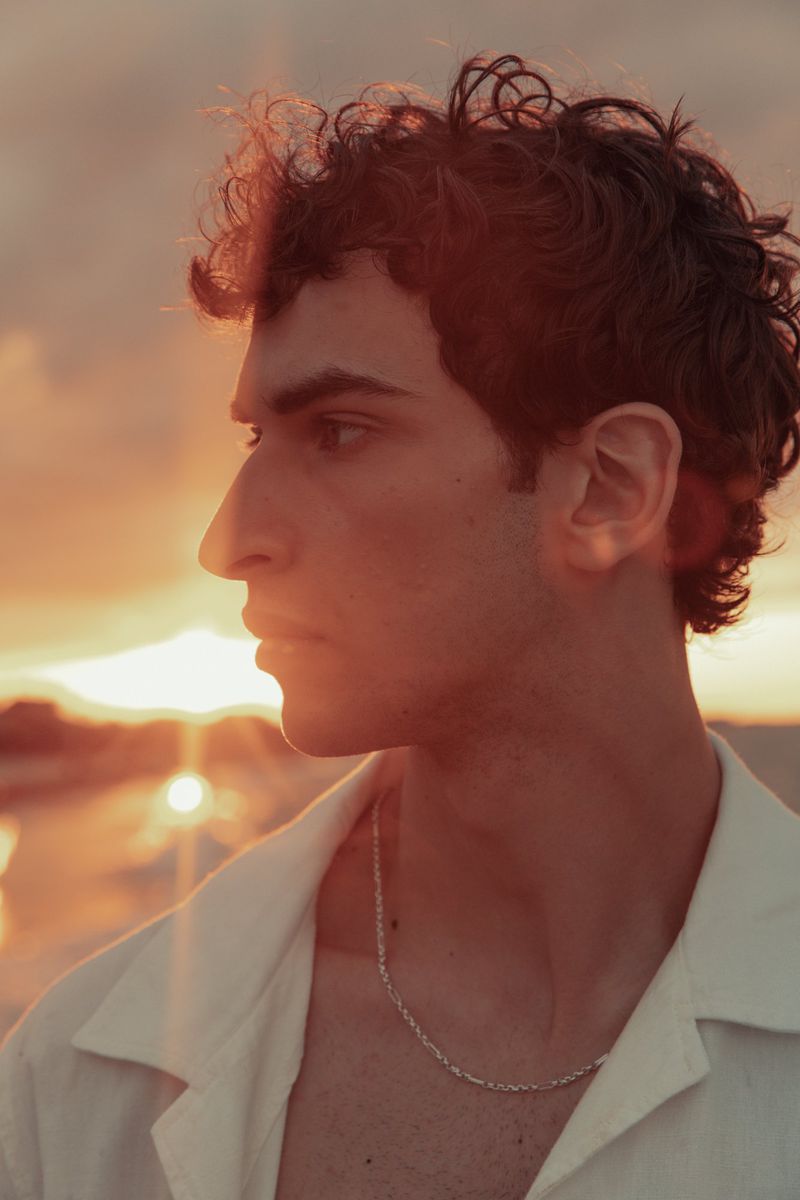 Portrait of an adult male with curly hair in profile, wearing a white shirt, against a sunset sky with warm orange and yellow tones.