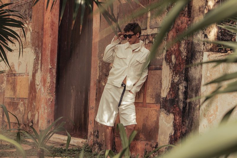 Adult male model poses in off-white jacket and shorts, accessorized with sunglasses and belt, leaning against aged wall near greenery outdoors.