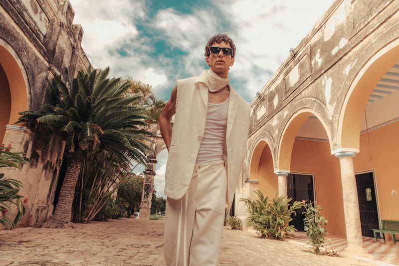 Man poses in off-white linen suit with sunglasses in a sunlit courtyard with palm trees and arched architecture.