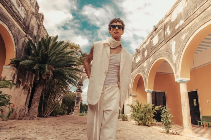 Man poses in off-white linen suit with sunglasses in a sunlit courtyard with palm trees and arched architecture.