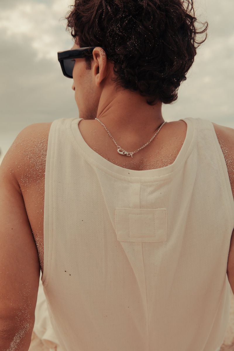 Man with dark hair and sunglasses wears a white tank top and silver necklace while standing on a sandy beach with an overcast sky.