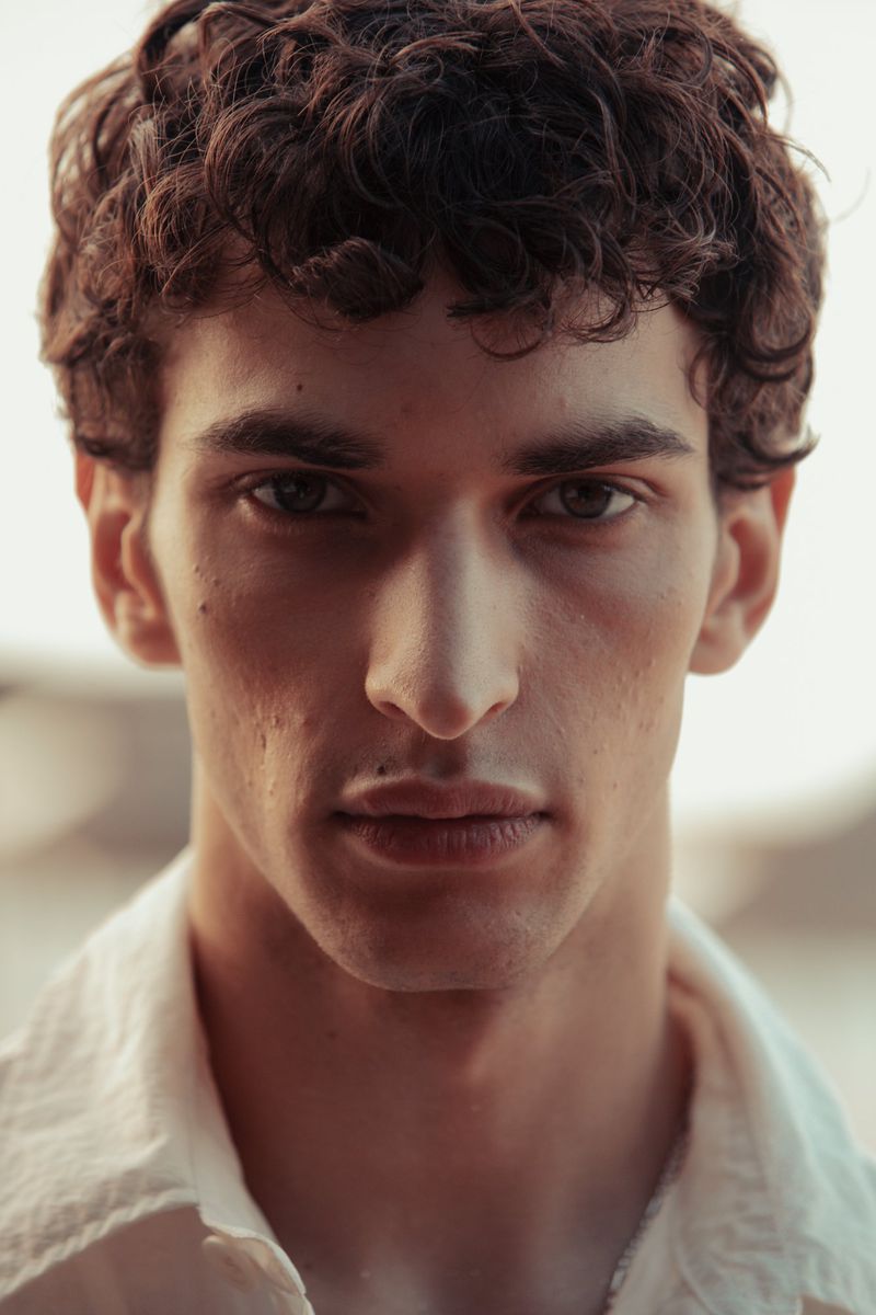 Close-up portrait of model Habib Masovic with curly brown hair in a white shirt for Zara Man S/S 24.