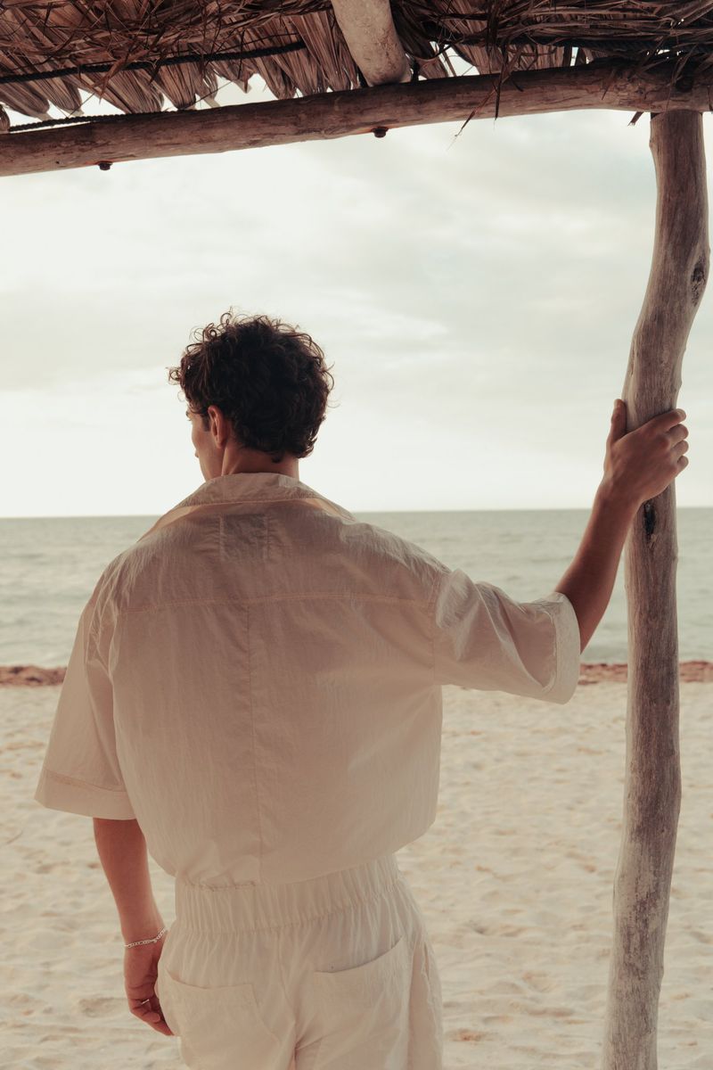 Person on beach looking at the ocean. Adult male with short brown hair wearing a short sleeved button down shirt and pants.