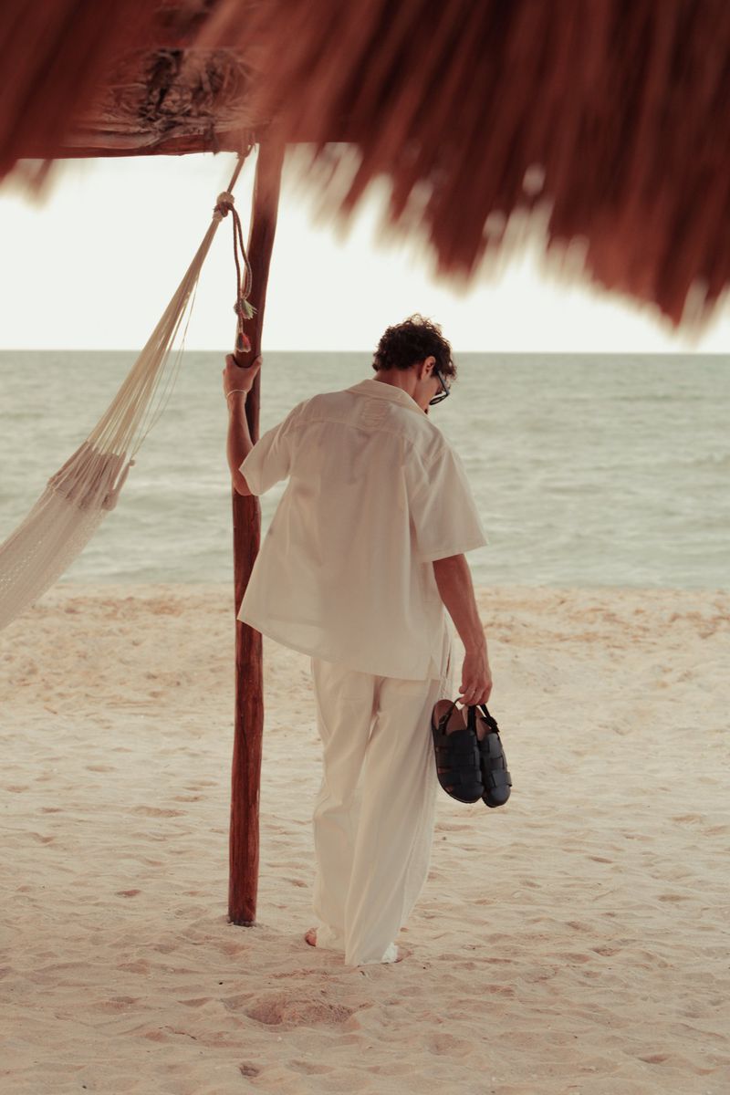 Back view of an adult male walking on a beach near the ocean and a hammock, holding a pair of shoes, wearing white clothing.
