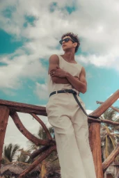 Model Habib Masovic wearing a cream sleeveless top and trousers while leaning against a wooden balcony railing under a blue sky.