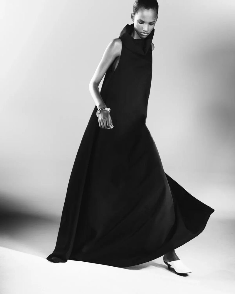 Model Edna Karibwami poses in a long sleeveless black maxi dress and white mule heels against a studio background.