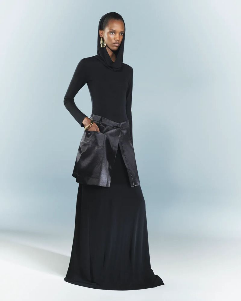 Full-length shot of an African American female fashion model in a black hooded top, long black skirt, and gold jewelry in a studio.