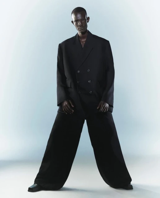 Fashion shot of a dark-skinned male model in an oversized black double-breasted suit and wide-leg pants against a gradient blue background.